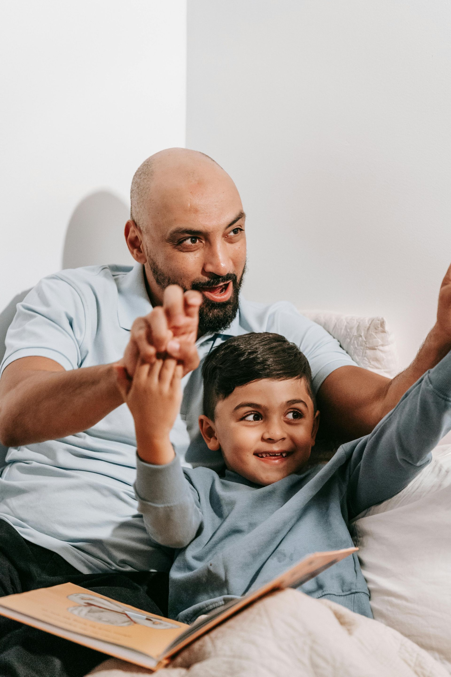 A man and a boy are sitting on a bed reading a book together.