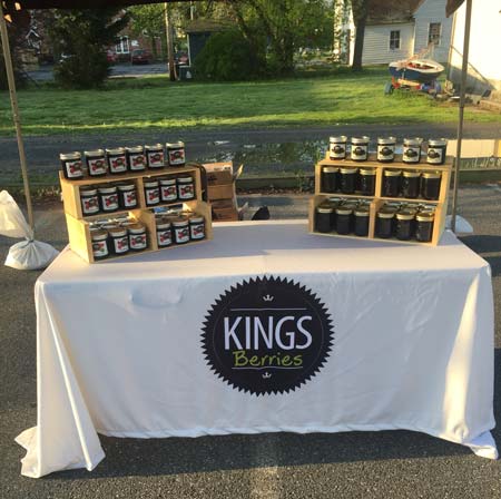 A white table with Kings Berries logo displays jars of jam on wooden risers at an outdoor market.