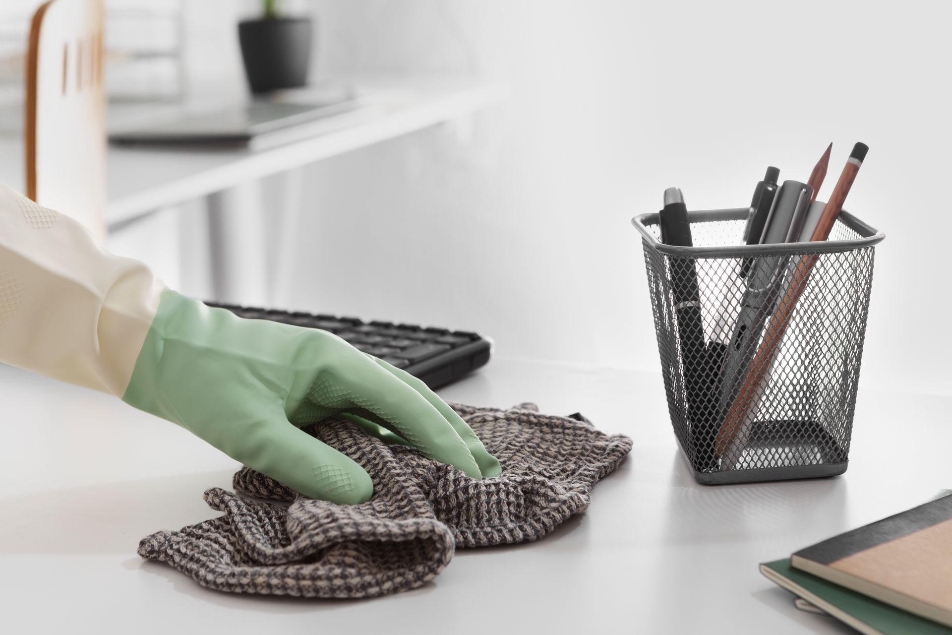 Person wearing green glove wiping a white desk with a cloth, near a pencil holder.