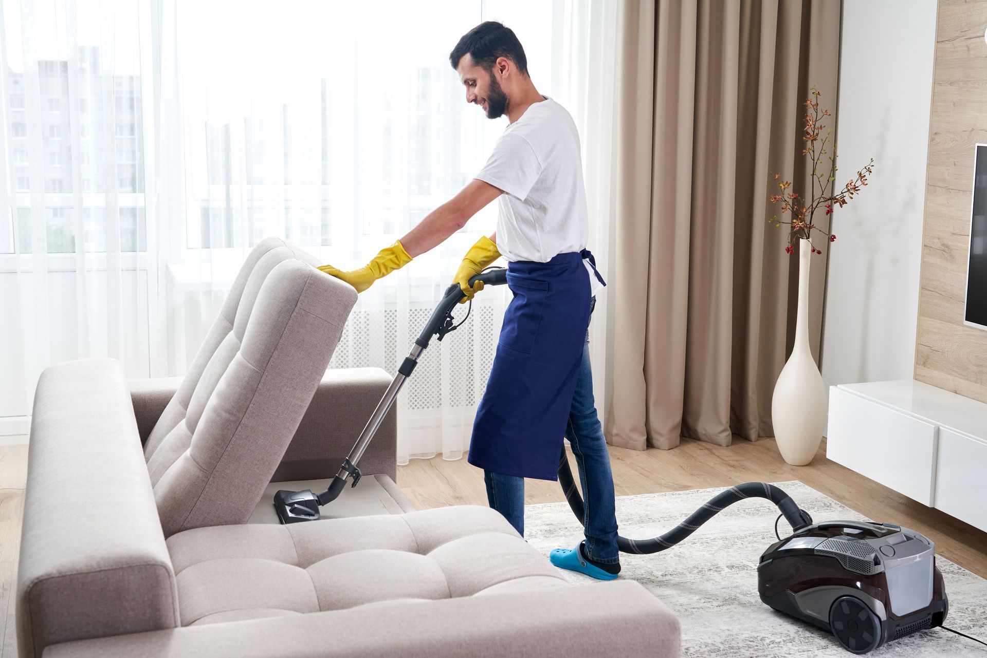 Man vacuums a beige sofa in a well-lit living room, wearing gloves and an apron.