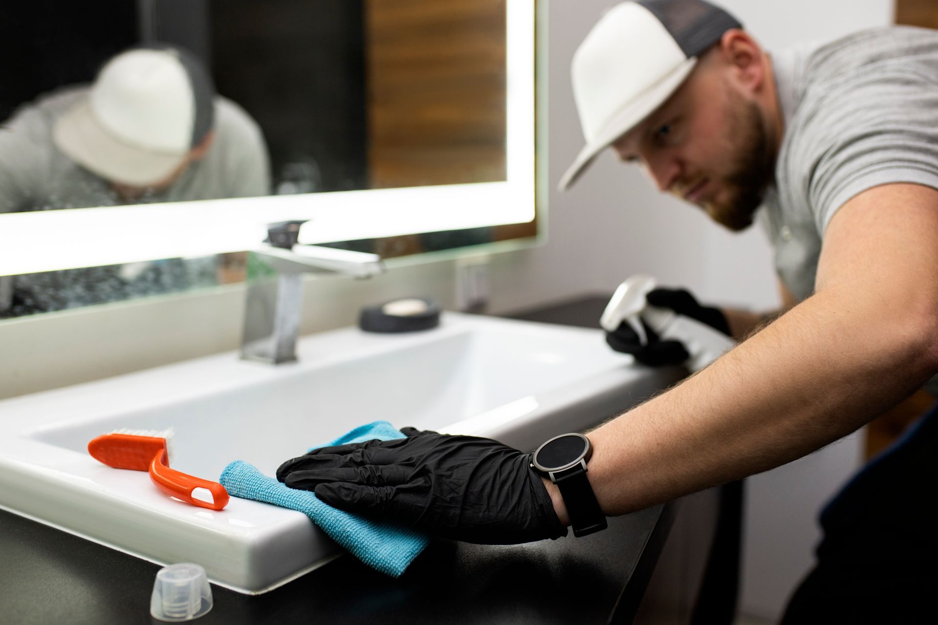 Person cleaning a white bathroom sink with a cloth, wearing gloves and a cap.