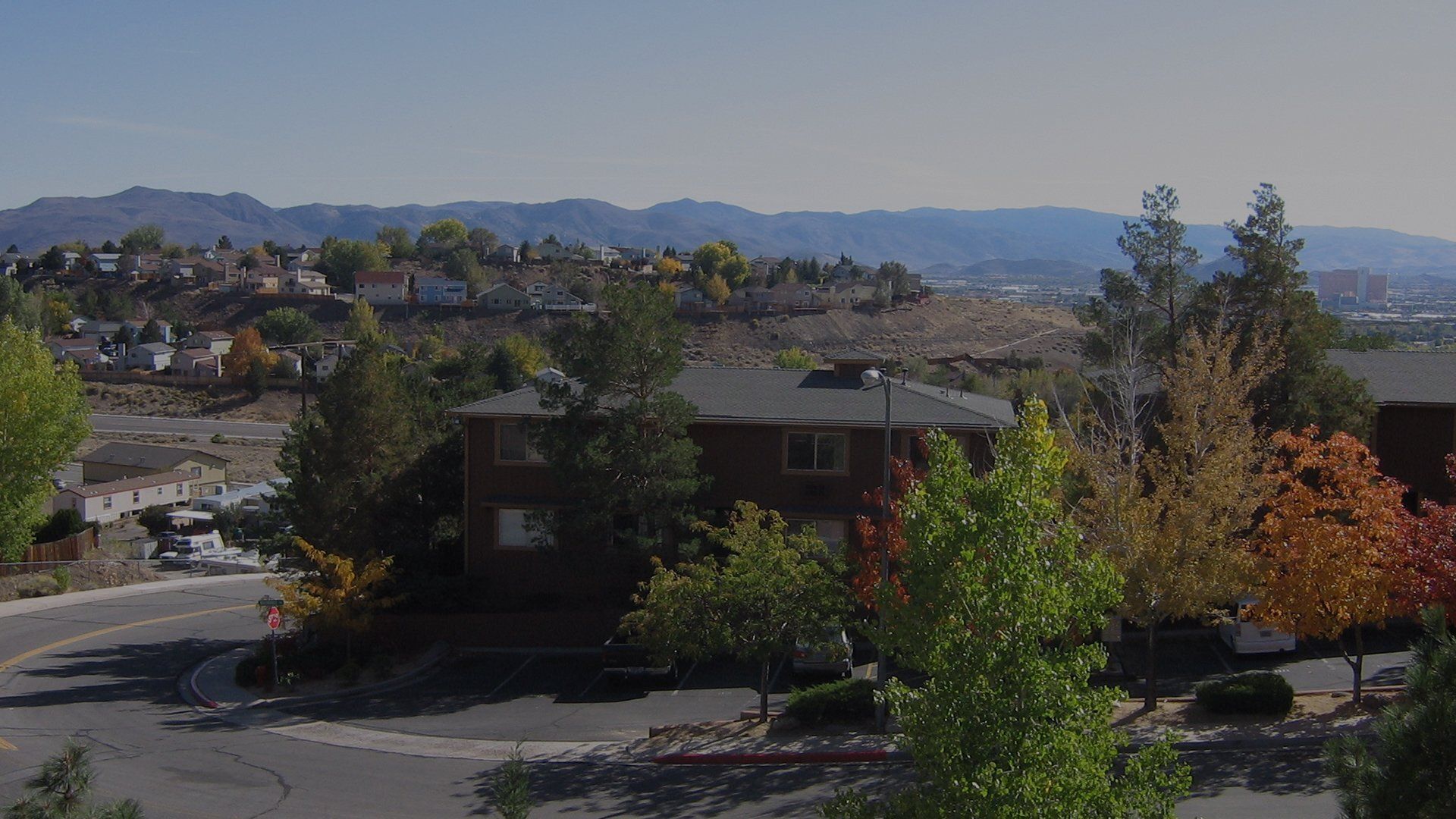 Street-level view of a hillside residential community with a brown building, trees, and distant mountains.