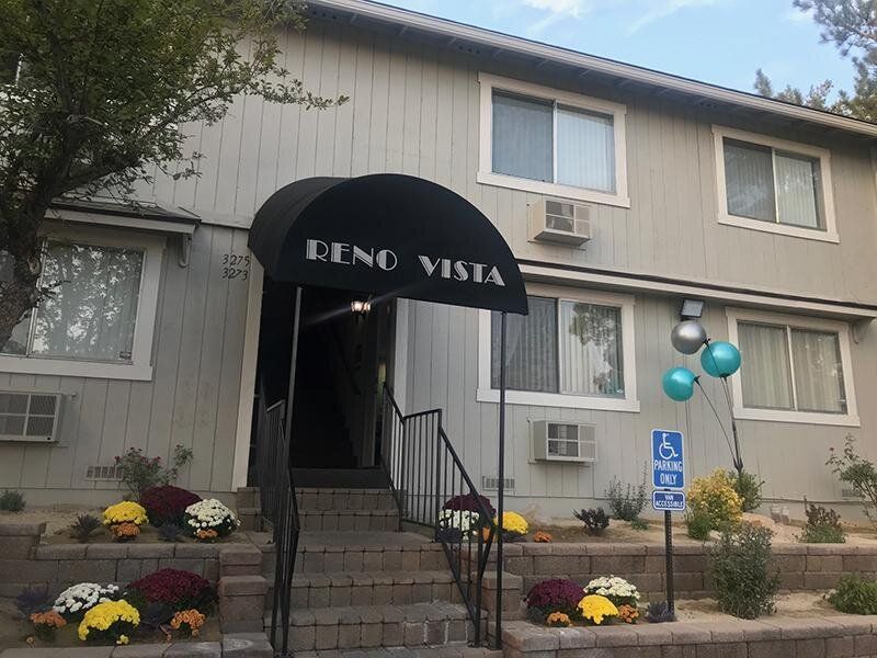 Exterior apartment building entrance with a curved black awning reading 'RENO VISTA,' stairs, and a handicap parking sign.