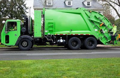 A green garbage truck is parked on the side of the road in front of a house.