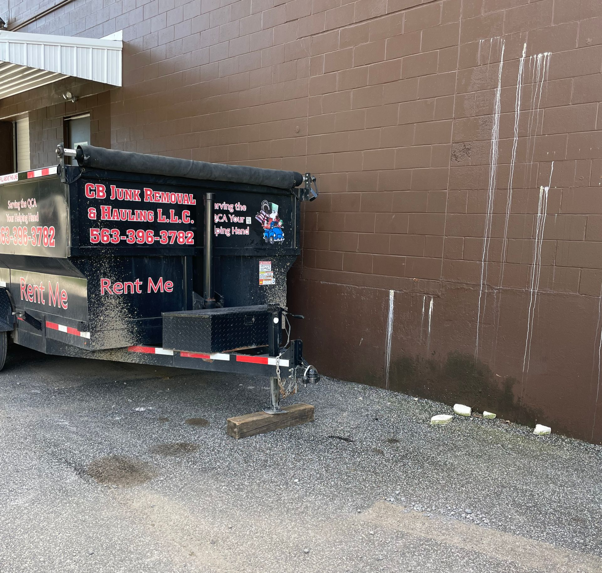 Dumpster trailer next to a brown brick wall. The trailer has text and is parked on gravel.