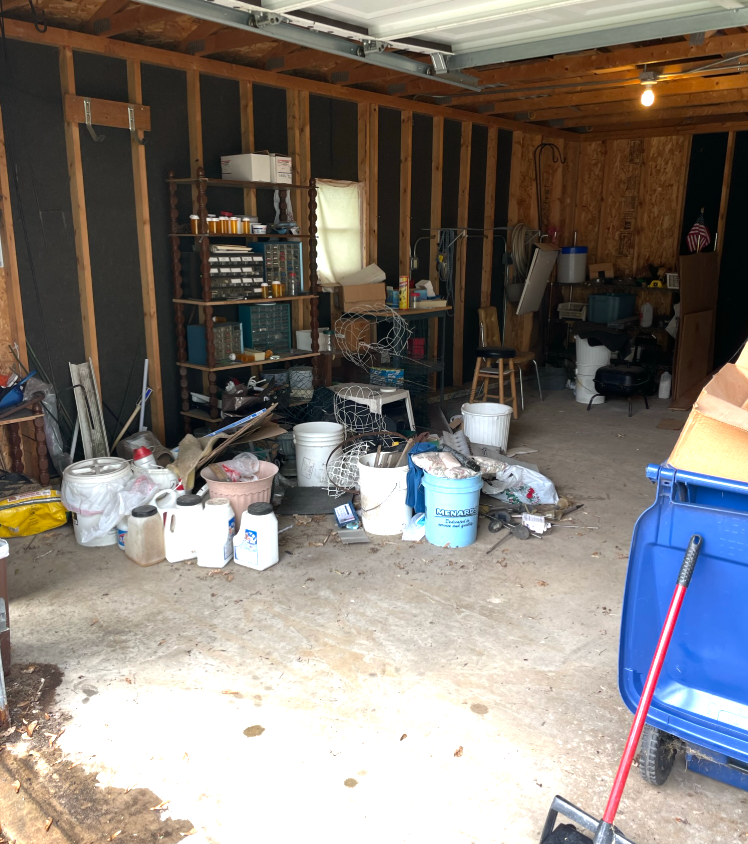 Cluttered garage interior; tools, shelves, and debris on floor. Blue wheelbarrow and white containers present.