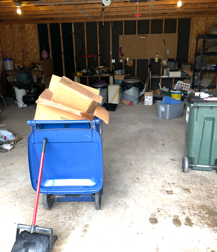 Messy garage with cardboard boxes in a blue bin, trash cans, and shelves holding various items.