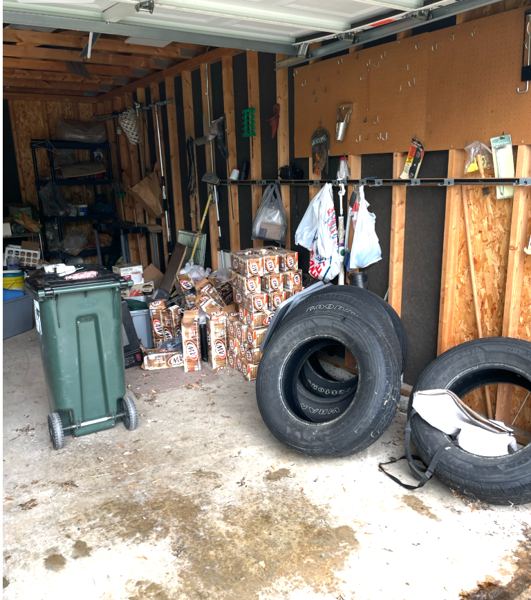 Cluttered garage interior: tires, boxes, trash can, tools, and general disarray visible against wooden walls.