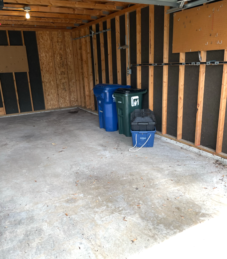 Empty garage with concrete floor, garbage bins along the back wall, and exposed wooden framing.