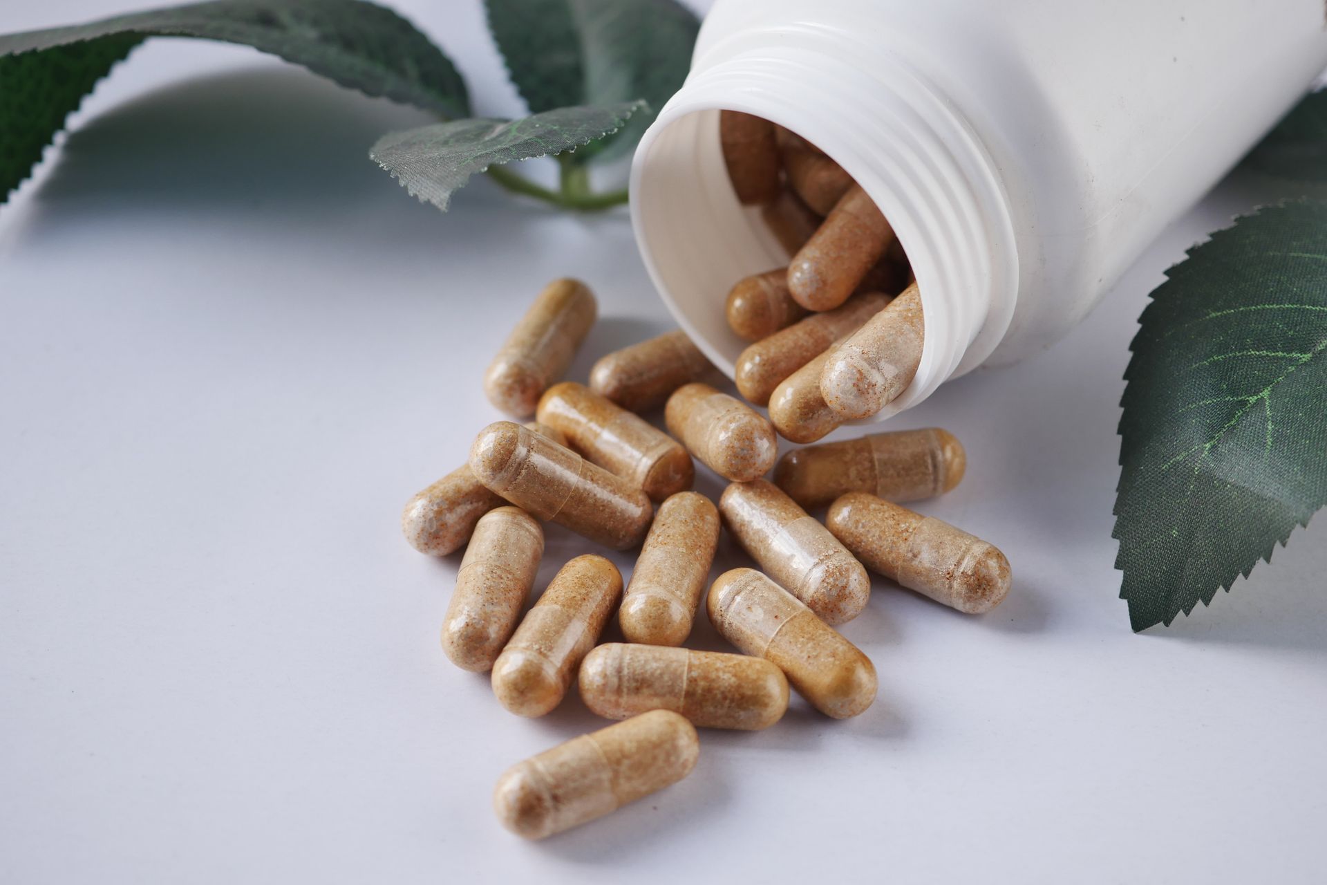 Beige capsules spilling from a white bottle, with green leaves in the background. Beige capsules spilling from a white bottle, with green leaves in the background.