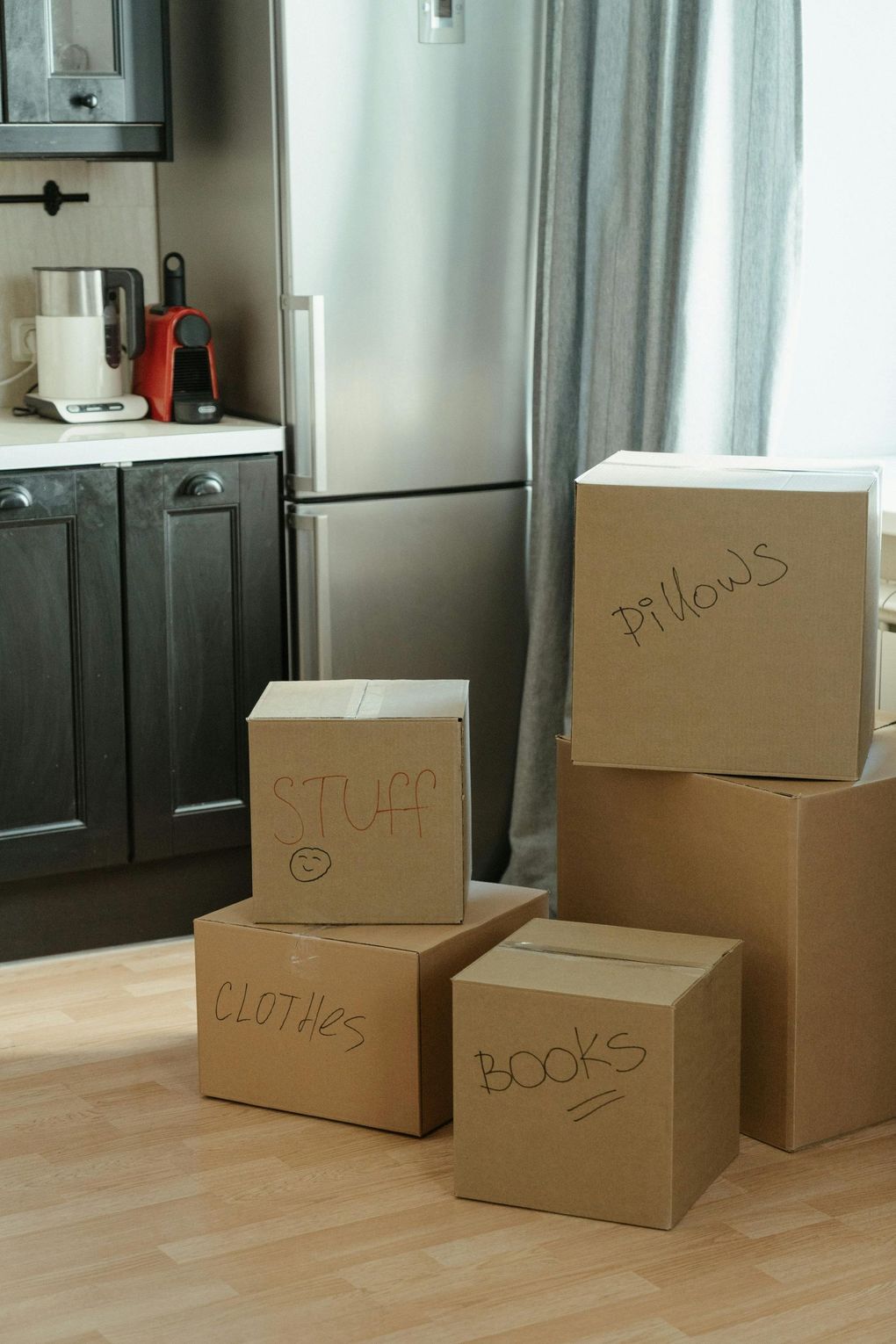Several cardboard boxes stacked on a kitchen floor beside a stainless steel refrigerator and cabinets