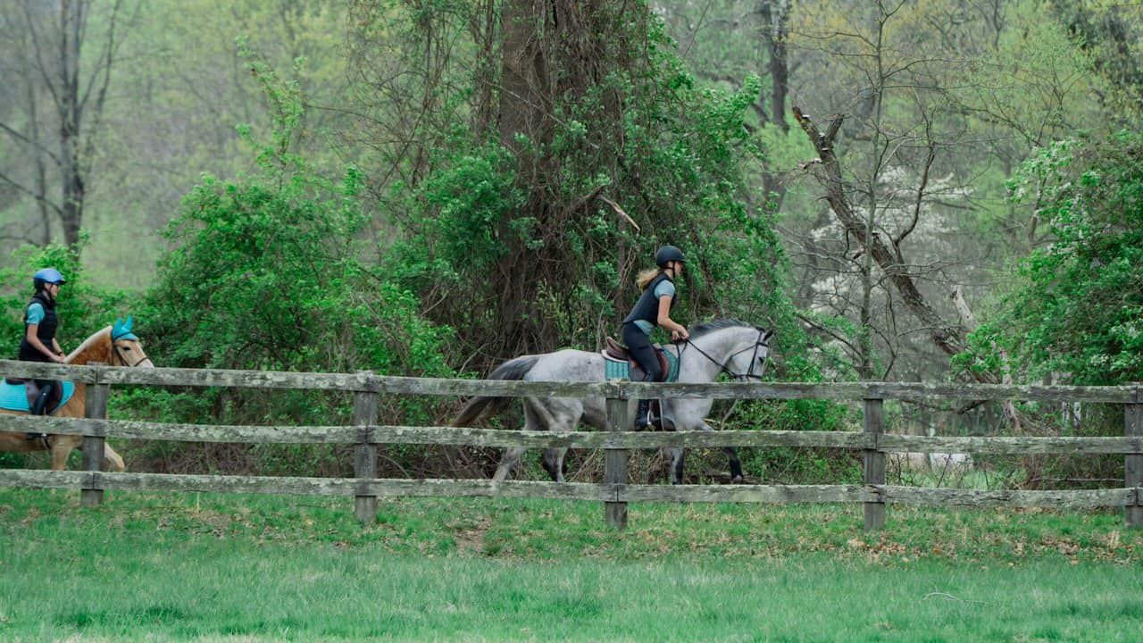 Two people are riding horses in a field behind a wooden fence.