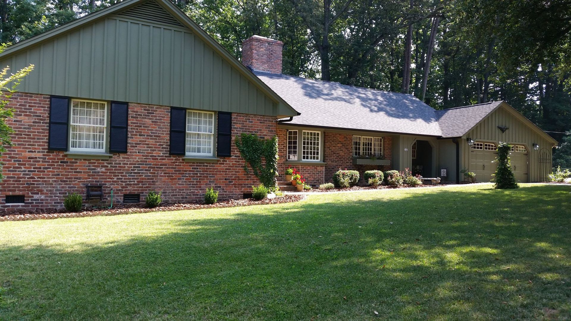 A sprawling single-story ranch-style home featuring red brick siding, olive-green vertical paneling, and a grassy yard.