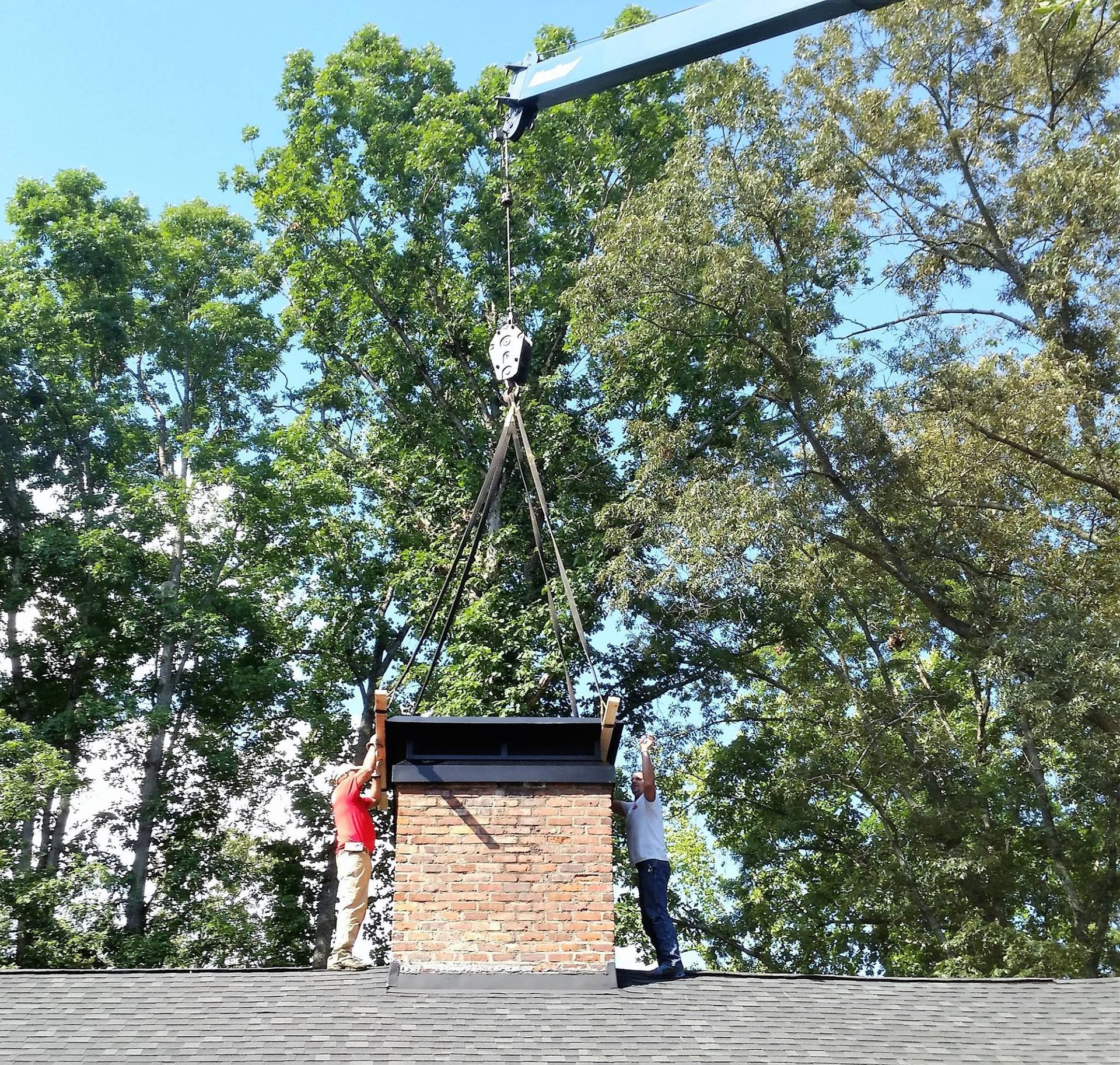 Two workers guide a brick chimney structure being lowered onto a roof by a crane against a backdrop of trees.