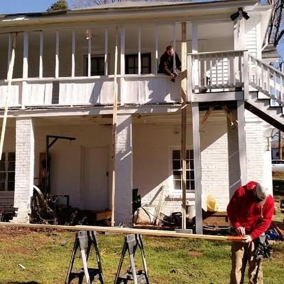 Two people work on a white two-story house renovation, with one person on the porch and another preparing lumber outside.