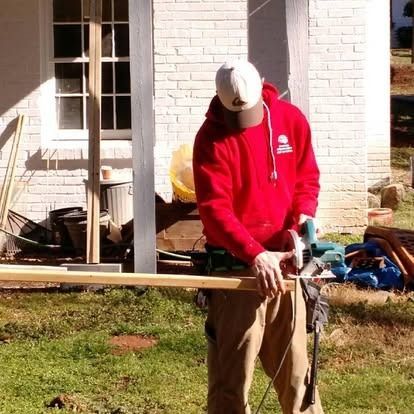 A person in a red hoodie and white cap uses a handheld circular saw to cut a wooden board outdoors near a white brick wall.