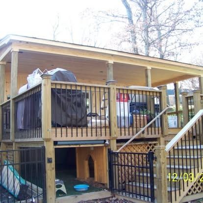 A two-story wooden deck with a covered upper patio, black metal railings, a staircase, and a ground-level pet enclosure.