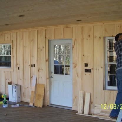 A person works on a house exterior featuring light-colored vertical wood siding, a white door, and windows on a porch.