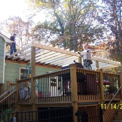 Two people work on a wooden pergola frame attached to the exterior of a green house above a raised deck.