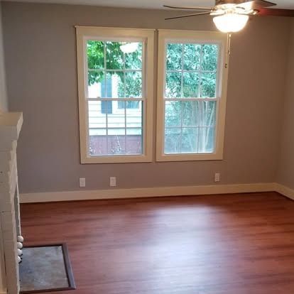 An interior view of a room with light grey walls, two windows, wood-look flooring, and a portion of a white fireplace.