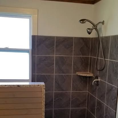 A shower area with dark gray square tiles, a silver dual shower head, a small corner shelf, and a partial view of a window.