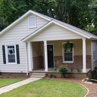 A single-story, beige-sided house with a covered front porch, brick foundation, dark shutters, and a black front door.