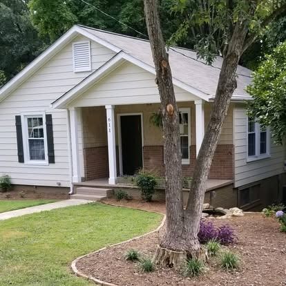 A light-colored, single-story suburban house with a front porch, tan siding, and dark shutters, viewed from the lawn.