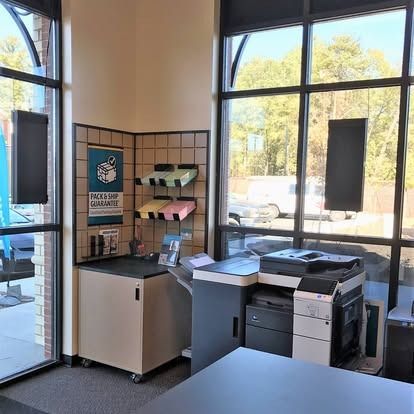A corner of an office with a copier, a small cabinet with paper slots, and a sign on a tiled wall next to large windows.