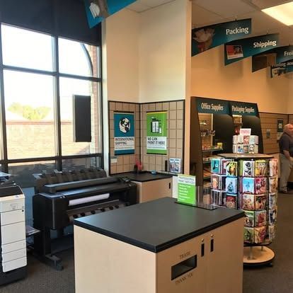 A print and ship center featuring a large printer, a service counter, office supplies, and a spinning greeting card rack.