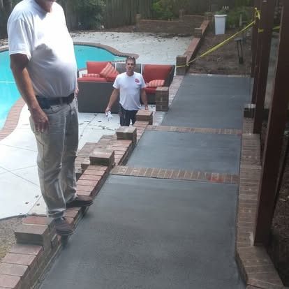 Two people stand on a newly poured concrete walkway with brick accents next to a backyard pool and patio area.