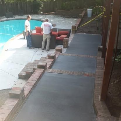 Two people standing on a patio next to a swimming pool and a new concrete walkway with brick borders.