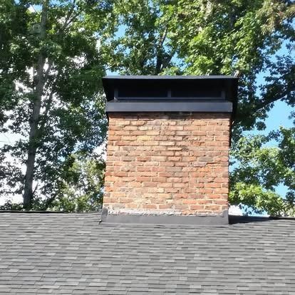 A brick chimney with a black metal cap, centered on a shingled roof against a background of green trees and blue sky.
