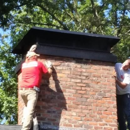 Two people stand on a roof, working on the black metal cap of a red brick chimney outdoors.