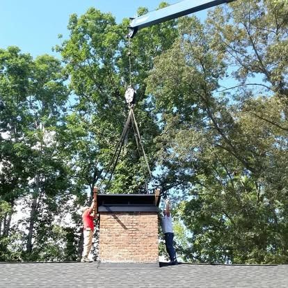 Two workers guide a brick chimney structure being lowered onto a roof by a crane.