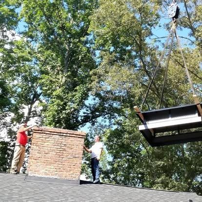 Two workers on a shingled roof prepare to install a large structural component being lowered by a crane.