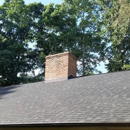 A brick chimney rising from a grey shingled roof against a background of green trees.