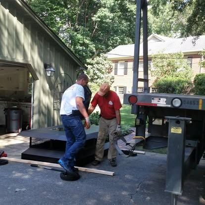 Two workers in work clothes stand on a driveway near a house, preparing to move a large, flat metal object.