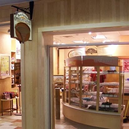 A chocolate shop store front with a hanging wooden sign, display cases filled with sweets, and light-toned wood walls.