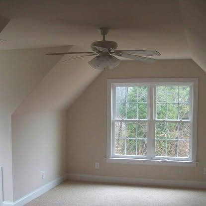 An empty room with beige walls, light carpet, a window with a tree view, and a ceiling fan.