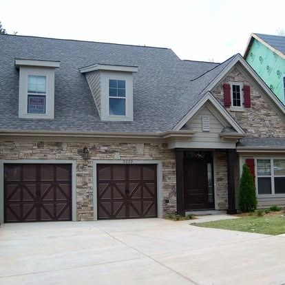A two-story house with stone veneer, grey shingled roof, two dark garage doors, and a small front yard.