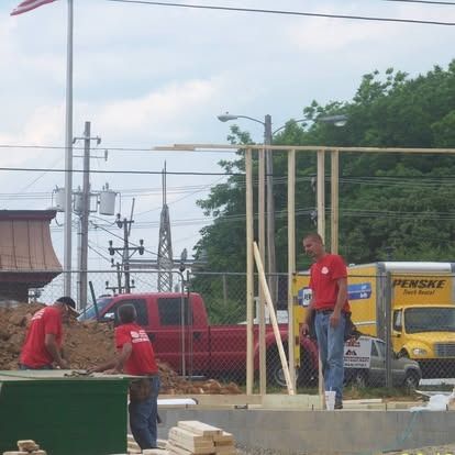 Two construction workers in red shirts work on a wooden wall frame at an outdoor job site near a red truck and Penske van.