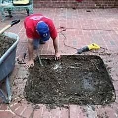 A person wearing a red shirt and blue cap kneels on a brick patio, digging into a square cutout in the ground.