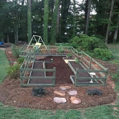 A U-shaped raised garden bed enclosed by a wooden fence, surrounded by mulch and stone path, set against a wooded backdrop.