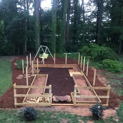 A backyard garden area with raised wooden beds, mulch paths, and wooden fence posts, set against a backdrop of trees.