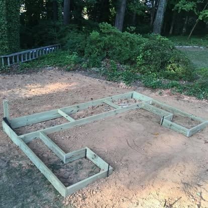 A light-colored wooden frame for a small building foundation sits on a level patch of dirt in a wooded area.