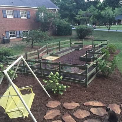 A fenced-in garden with raised beds in a backyard, featuring a yellow swing, stone path, and a small cat in the foreground.