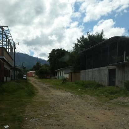 A dirt road stretches through a residential area with simple buildings, trees, and distant mountains under a cloudy sky.