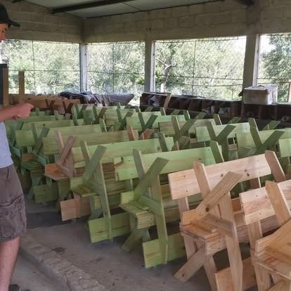 Rows of unfinished wooden benches arranged in a rustic, open-air workshop with a person partially visible in the foreground.