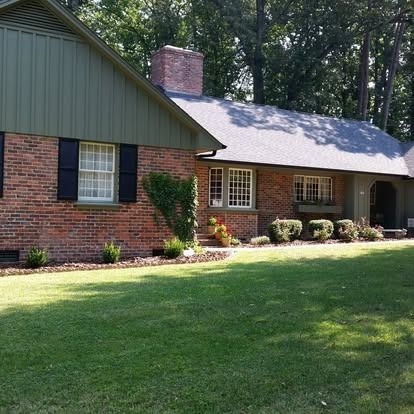 Single-story brick house with a dark green gabled roof and siding, surrounded by a green lawn and mature trees.