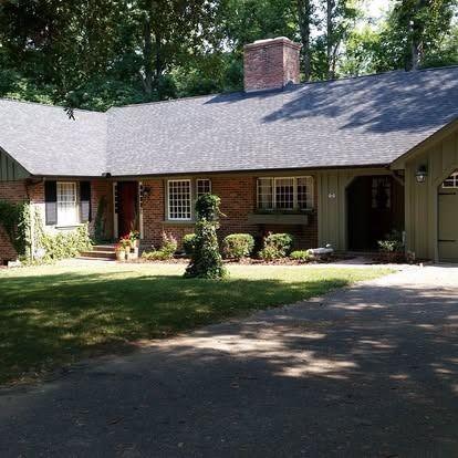 A single-story brick house with a dark shingled roof, arched entrance, and lush green landscaping on a sunny day.